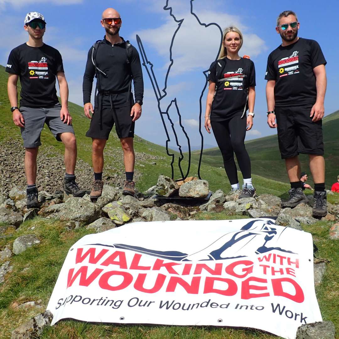 Four people in hiking gear stand on a rocky hilltop next to a metal figure sculpture, with a "Walking with the Wounded" banner in front.