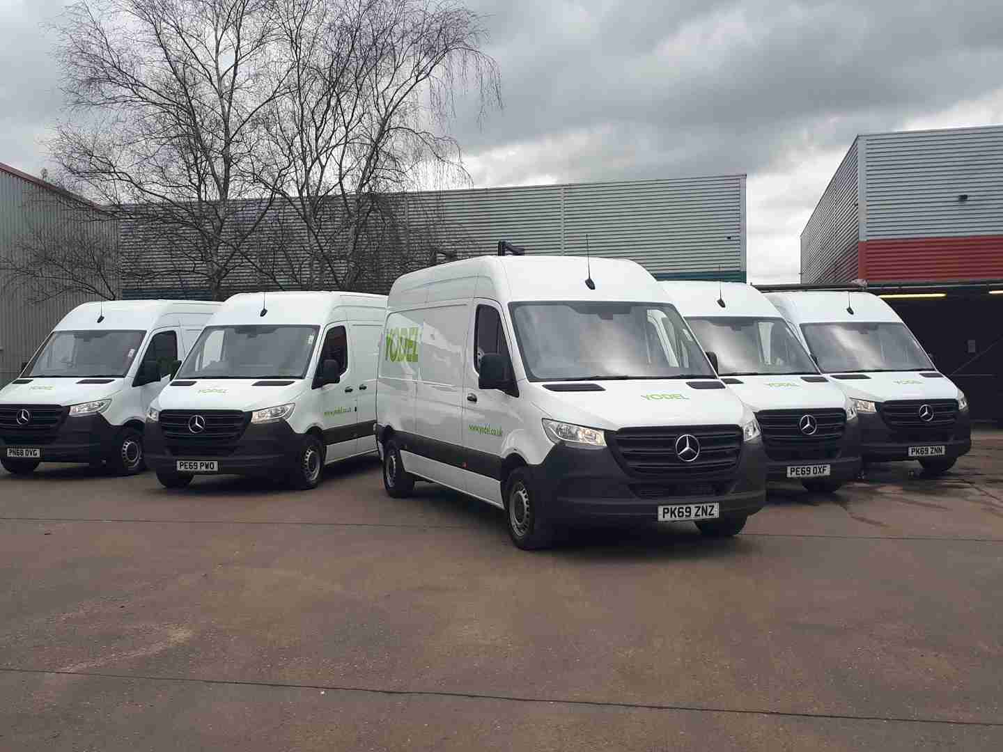 Five white delivery vans with Yodel logo parked in a row on a concrete lot, with industrial buildings and a leafless tree in the background.