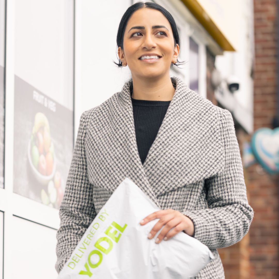Smiling woman in a gray coat holds a Yodel package outside a store, with a fruit poster visible in the background.